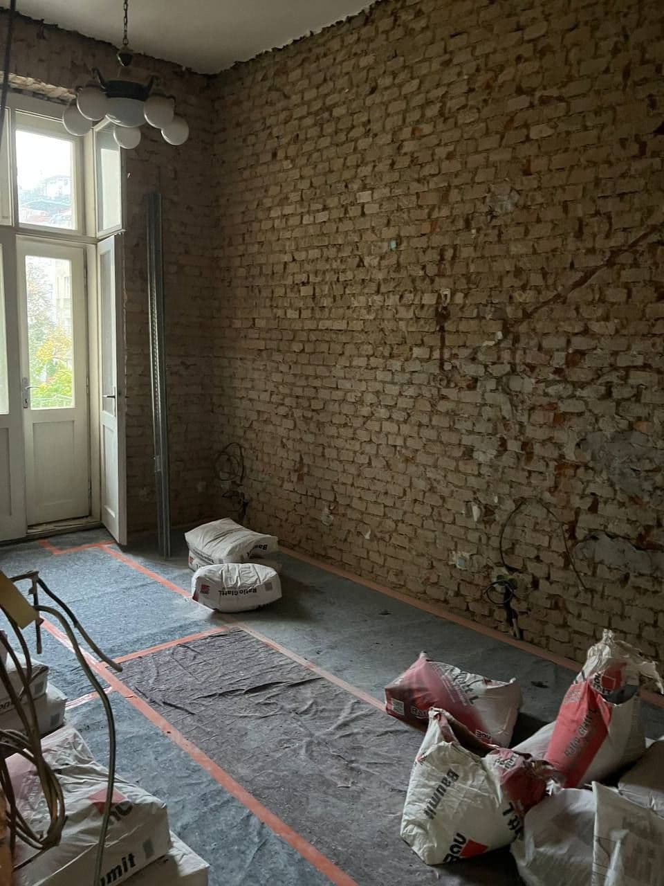 Pokoj vybouraný až na cihelné zdivo, balkonové dveře a podlaha zakrytá ochrannou textilií.